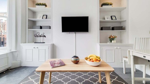 Ein modernes Wohnzimmer mit Fernseher, weißen Schränken, einem Esstisch mit Obstschale und einer Zeitschrift.
