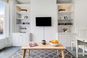 Een moderne woonkamer met een televisie, witte kasten, een eettafel met een fruitschaal en een tijdschrift.