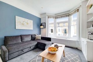 Een moderne woonkamer met een grijze bank, een houten salontafel met fruit, een blauwe accentmuur en grote ramen.