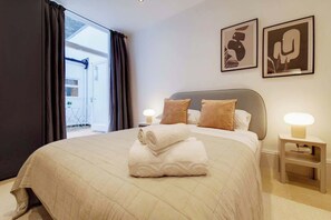 Una habitación de hotel con cama, dos lámparas de noche, una ventana con cortinas y dos cuadros en la pared.