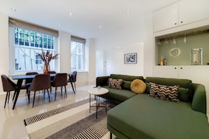 Ein modernes Wohnzimmer mit einem grünen Sofa, einem Esstisch mit Stühlen, einem kleinen Beistelltisch und einem großen Fenster mit Blick ins Grüne.