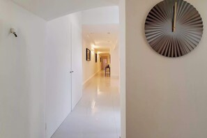 Un pasillo con una puerta blanca, un reloj en la pared y una mesita con un jarrón.