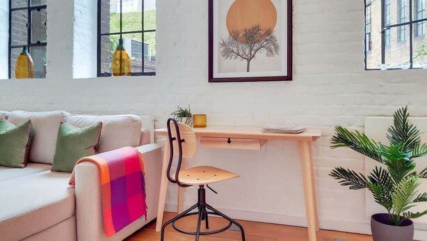 A room with a white brick wall, a desk with a chair, a sofa with cushions, a framed picture, and a potted plant. - VY6 - Stylish Loft-Style 1-Bed Near Borough Market (Greater London)