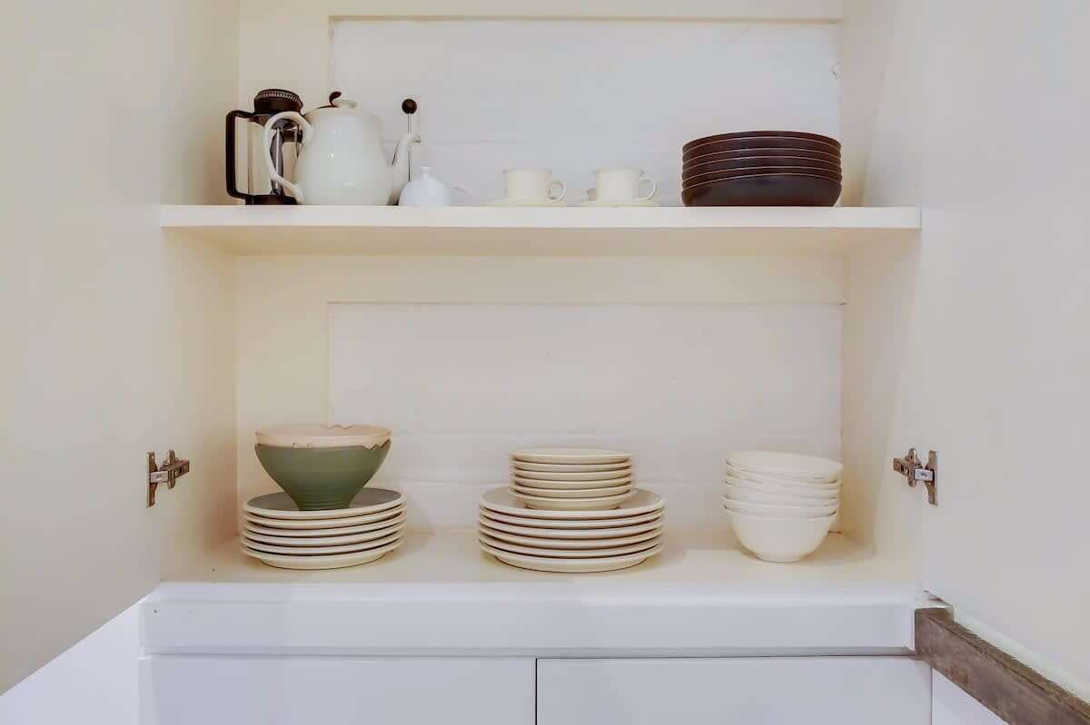 A kitchen with white cabinets, a white countertop, and a shelf with kitchenware.