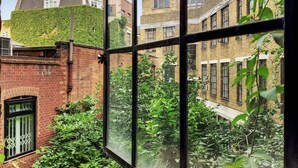 A view through a window with a brick building, a green ivy-covered wall, and a row of windows. - VY6 - Stylish Loft-Style 1-Bed Near Borough Market (Greater London)