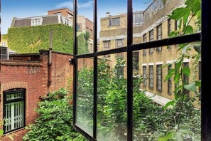 Blick durch ein Fenster auf ein Ziegelgebäude, eine mit Efeu bewachsene grüne Mauer und eine Reihe von Fenstern.