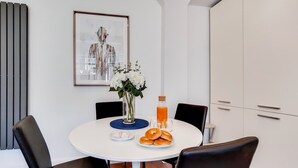 Un comedor con una mesa redonda, sillas negras, un jarrón con flores, una botella de jugo de naranja y un plato de bagels.