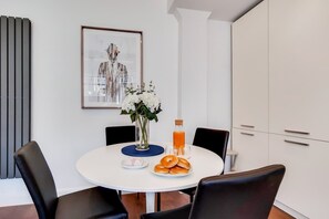 Ein Essbereich mit einem runden Tisch, schwarzen Stühlen, einer Blumenvase, einer Orangensaftflasche und einem Teller mit Beugeln.