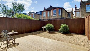 Un patio trasero con una terraza de madera, una mesa pequeña y dos plantas en macetas.