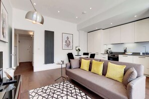 Ein modernes Wohnzimmer mit einer Couch, einem Couchtisch, einer Pendelleuchte und einer Küche im Hintergrund.