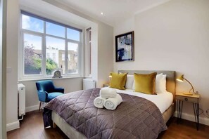 Ein Schlafzimmer mit großem Fenster, einem Bett mit Steppdecke, zwei Nachttischlampen, einem Sessel und einem Heizkörper.