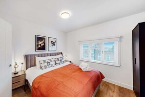 A hotel room with a bed, a nightstand, a lamp, a window with blinds, and artwork on the wall. - DA5 - Contemporary 2-Bedroom Duplex Penthouse (Greater London)