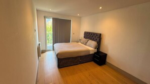 A bedroom with a bed, a bedside table, a nightstand, and a window with curtains. - The Zenith Residence - 2 Bedroom (London)