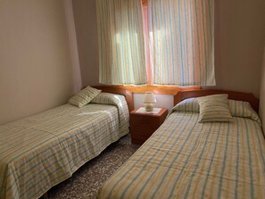 Ein Zimmer mit zwei Betten, einem Nachttisch mit Lampe und einem Fenster mit Vorhängen.