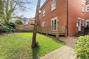 A brick building with a wooden deck, a grassy yard, and a tree. - Comfortable 1 BDR Flat in Winchester (Winchester)