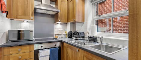 A modern kitchen with wooden cabinets, a stainless steel range hood, a microwave, a coffee maker, and a window with a brick wall view.