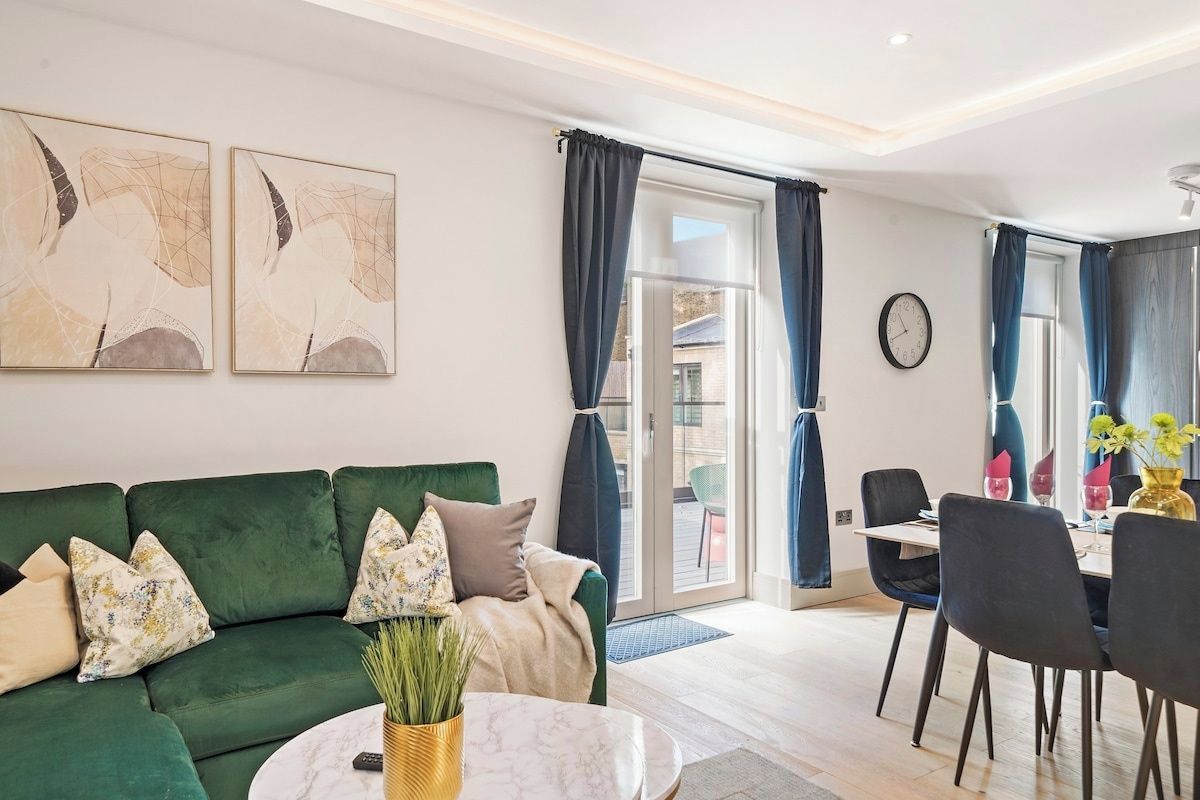 Ein modernes Wohnzimmer mit einem grünen Sofa, einem Essbereich mit Tisch und Stühlen sowie einem großen Fenster mit Vorhängen.