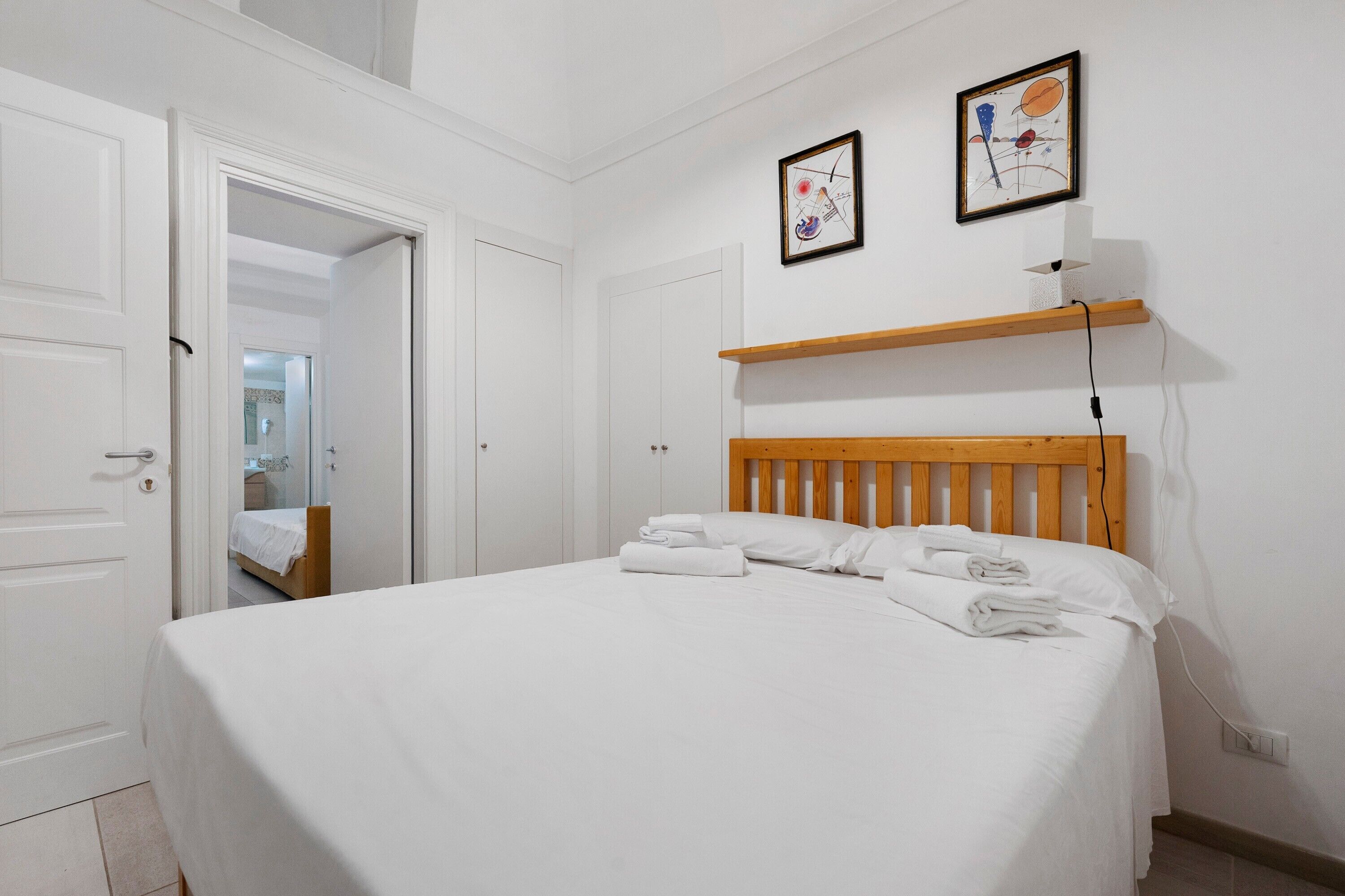 A bedroom with a bed, a wooden headboard, two framed artworks on the wall, and a shelf with a lamp.