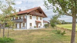 Une maison à deux étages, avec un toit recouvert de tuiles rouges, un balcon en bois et une cour verdoyante.