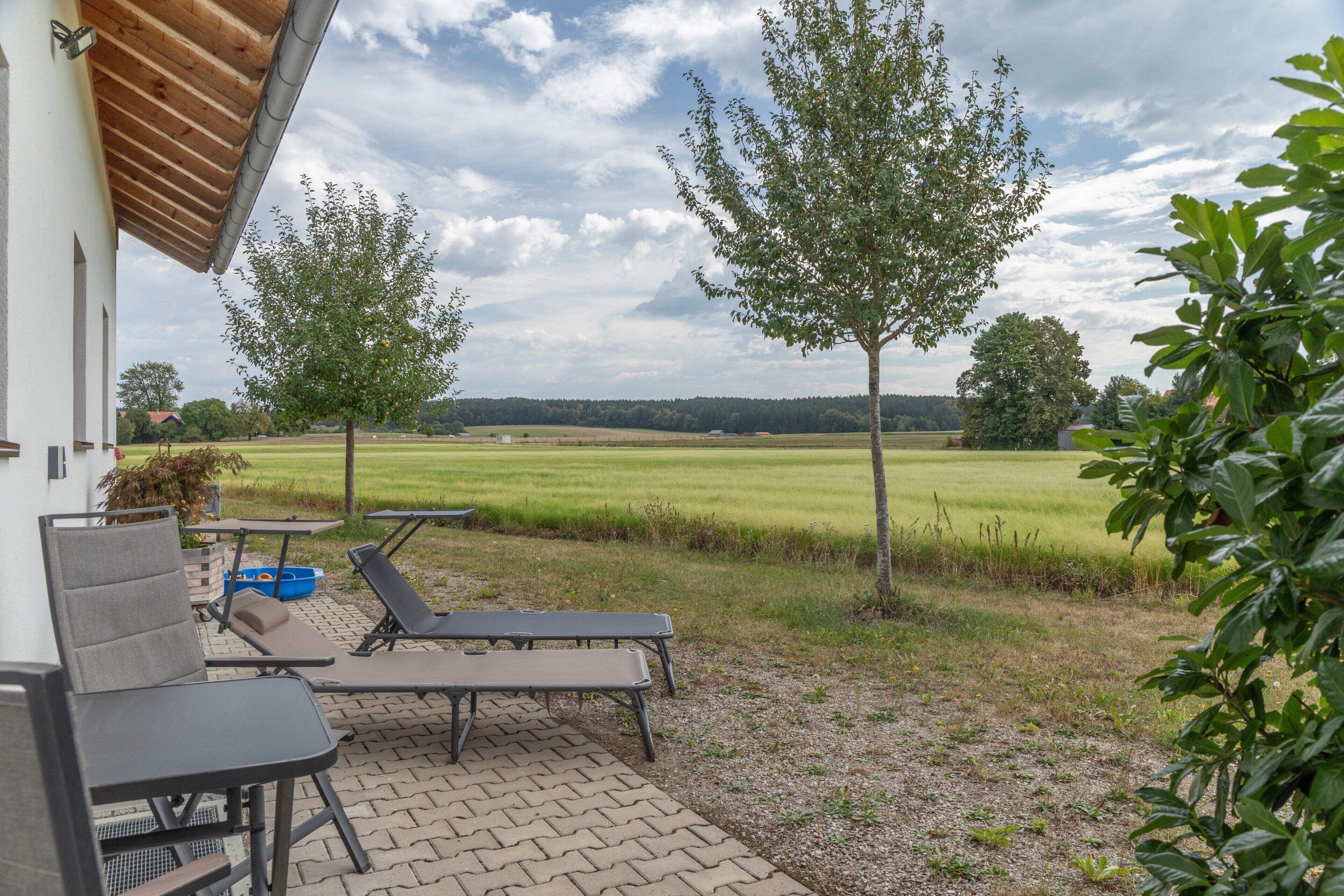 A patio with two lounge chairs and a table, overlooking a grassy field and trees.
