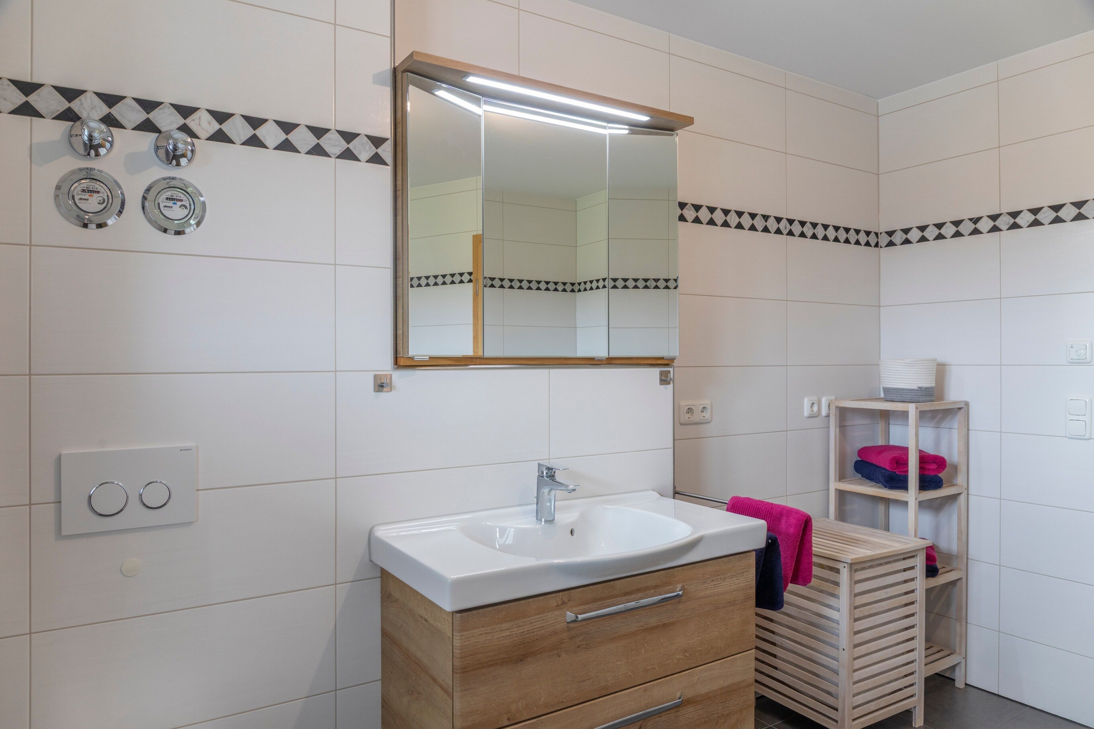 Un baño con un lavabo blanco, tocador de madera, un espejo y un perchero con toallas dobladas.
