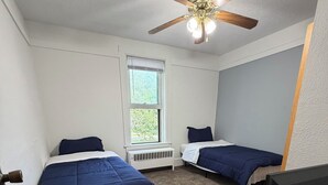 A room with two beds, a ceiling fan, a window, a desk with a computer, and a trash bin.