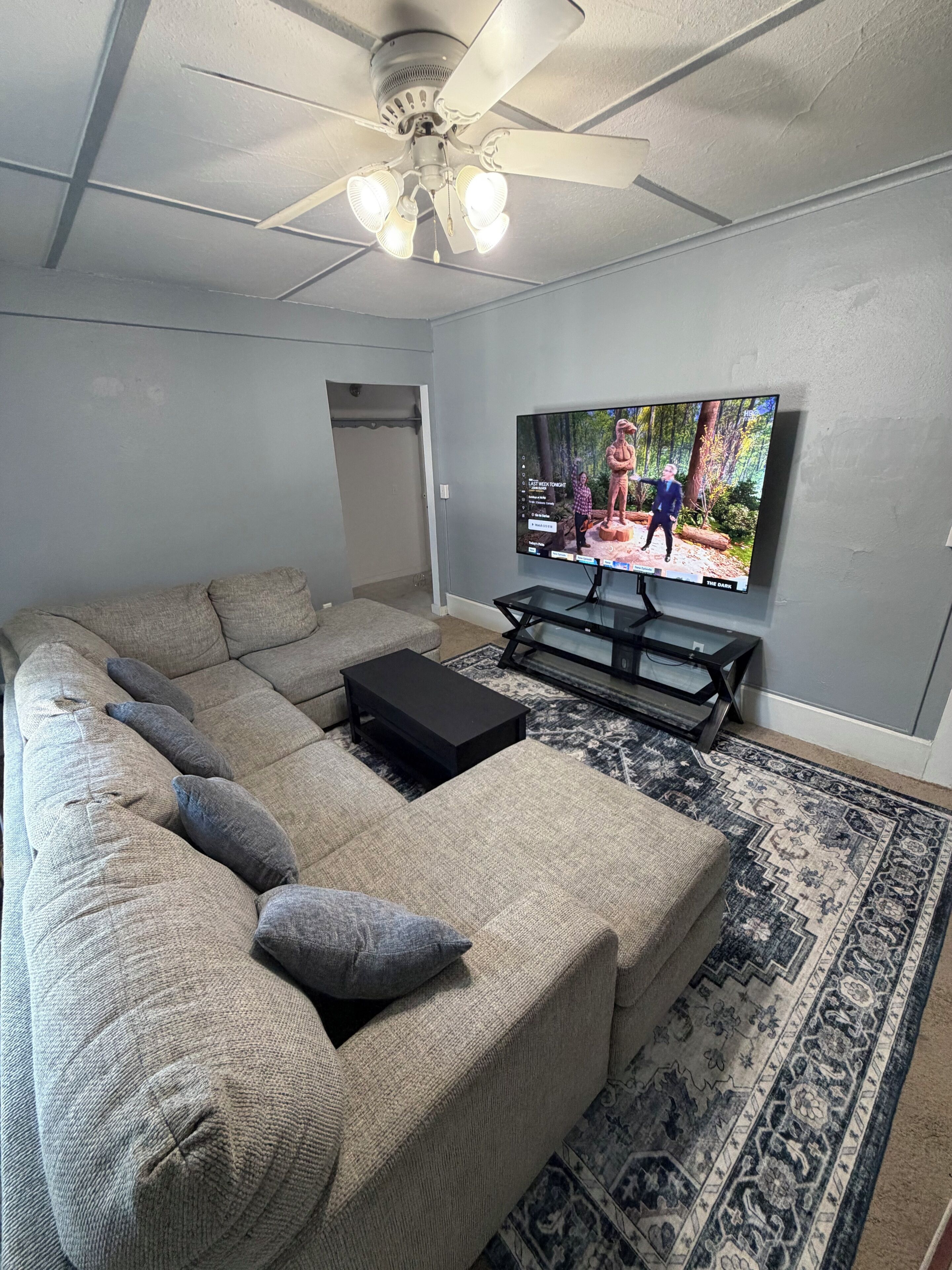 Un salon comprenant un canapé d’angle, un téléviseur à écran plat, un ventilateur de plafond et un tapis à motifs.
