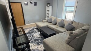 A living room with a beige sofa, a coffee table, a TV, a ceiling fan, and a window with blinds.