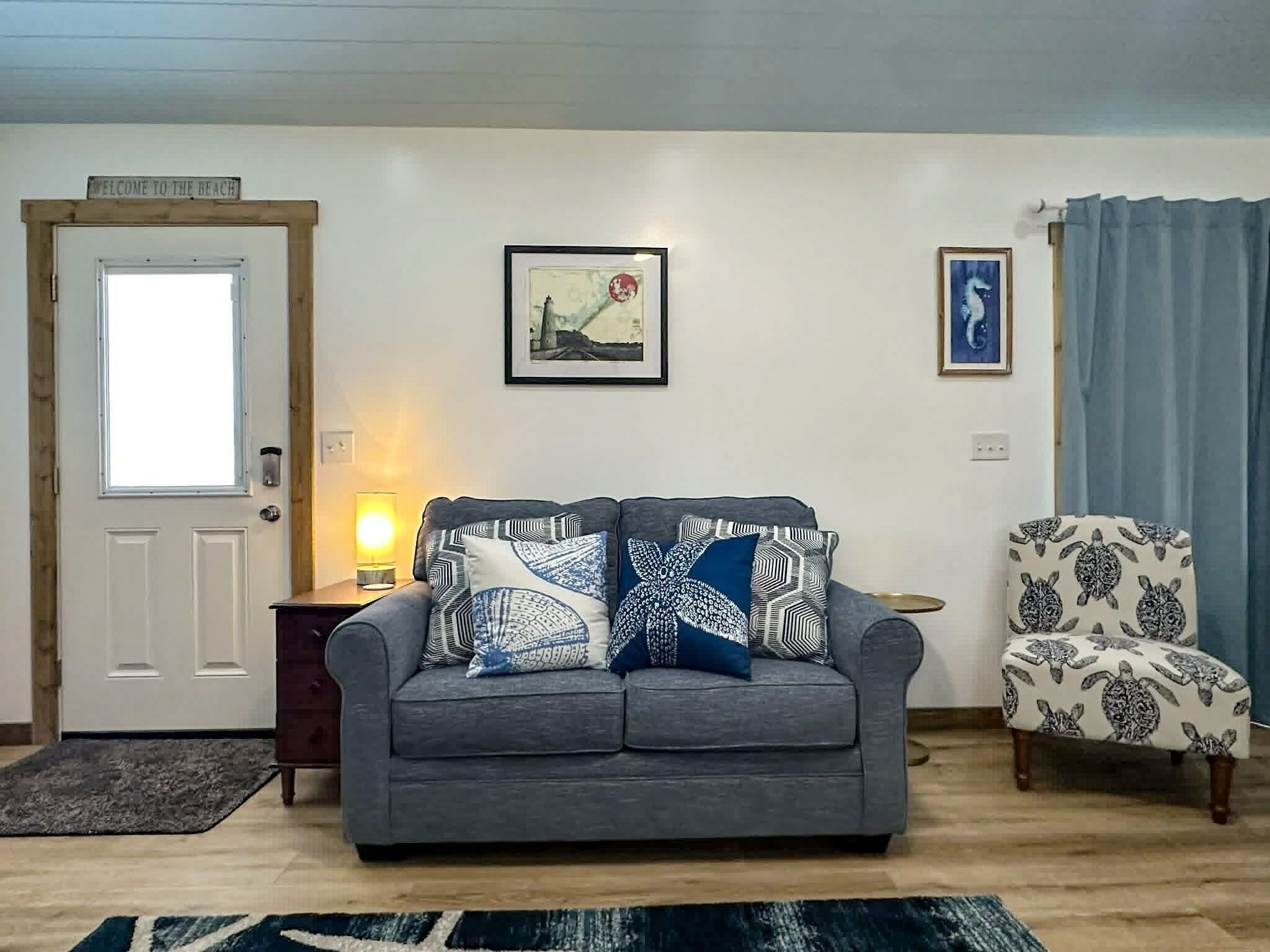Un soggiorno con un divano grigio, cuscini decorati, un quadro incorniciato, una poltrona con motivo a tartaruga e una porta con il cartello 'Welcome to the Beach'.