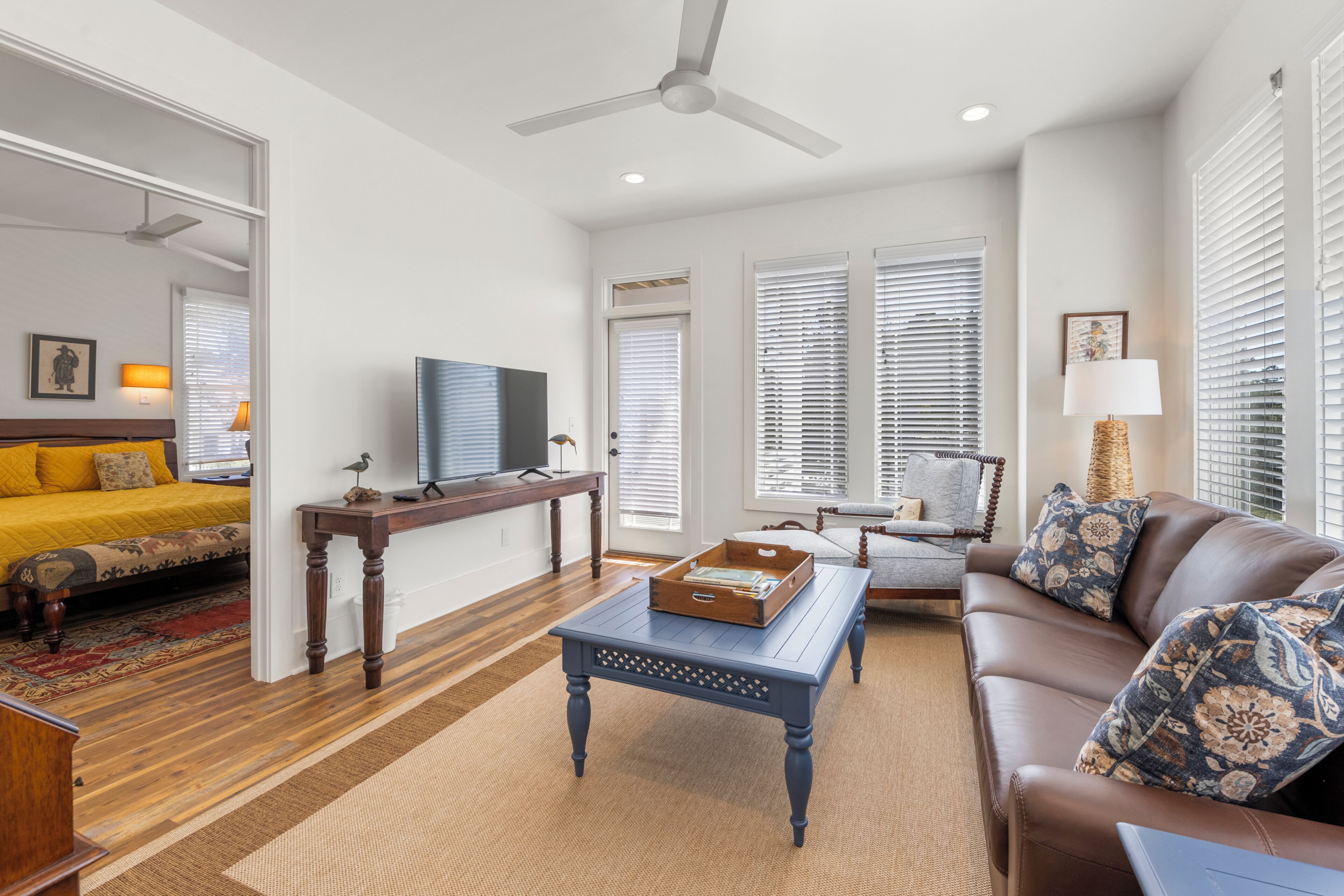 A modern living room with a sofa, coffee table, and a television. There is a bed with a yellow bedspread and a window with blinds.
