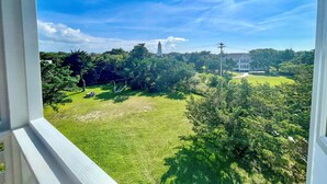 A view from a window overlooking a well-maintained lawn, a white building, and a lighthouse in the distance. - Wyf of Bathe: Silver Lake Views with docking available. (Ocracoke)