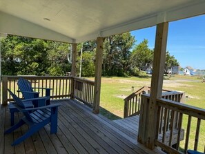 A wooden deck with a railing, a blue Adirondack chair, and a view of a grassy area with trees and houses in the distance. - Ocracoke Local 4: Harbor Views | Private Boat Slip | King Bed (Ocracoke)