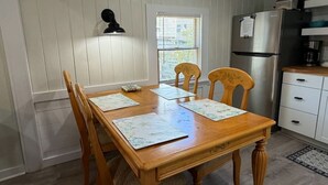 Un comedor con mesa de madera, cuatro sillas, una ventana con persianas, un refrigerador y un pequeño adorno en la pared.