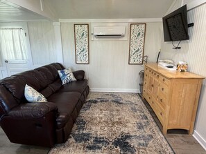 A living room with a brown leather sofa, a patterned rug, a wooden cabinet, and a mounted TV. - Ocracoke Local 4: Harbor Views | Private Boat Slip | King Bed (Ocracoke)