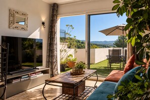 A modern living room with a flat-screen TV, a wooden coffee table, a sofa, and a view of a green landscape through large sliding glass doors. - Idylle apartment – Sleeps 2, with a large terrace and a view (Pertuis)