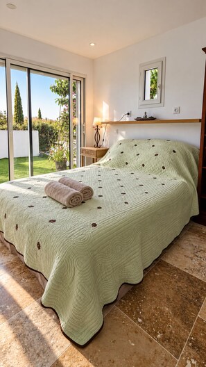 Ein Schlafzimmer mit einem großen Bett, einer Schiebetür zu einem Garten, einem Nachttisch mit Lampe und einem kleinen Regal über dem Bett.