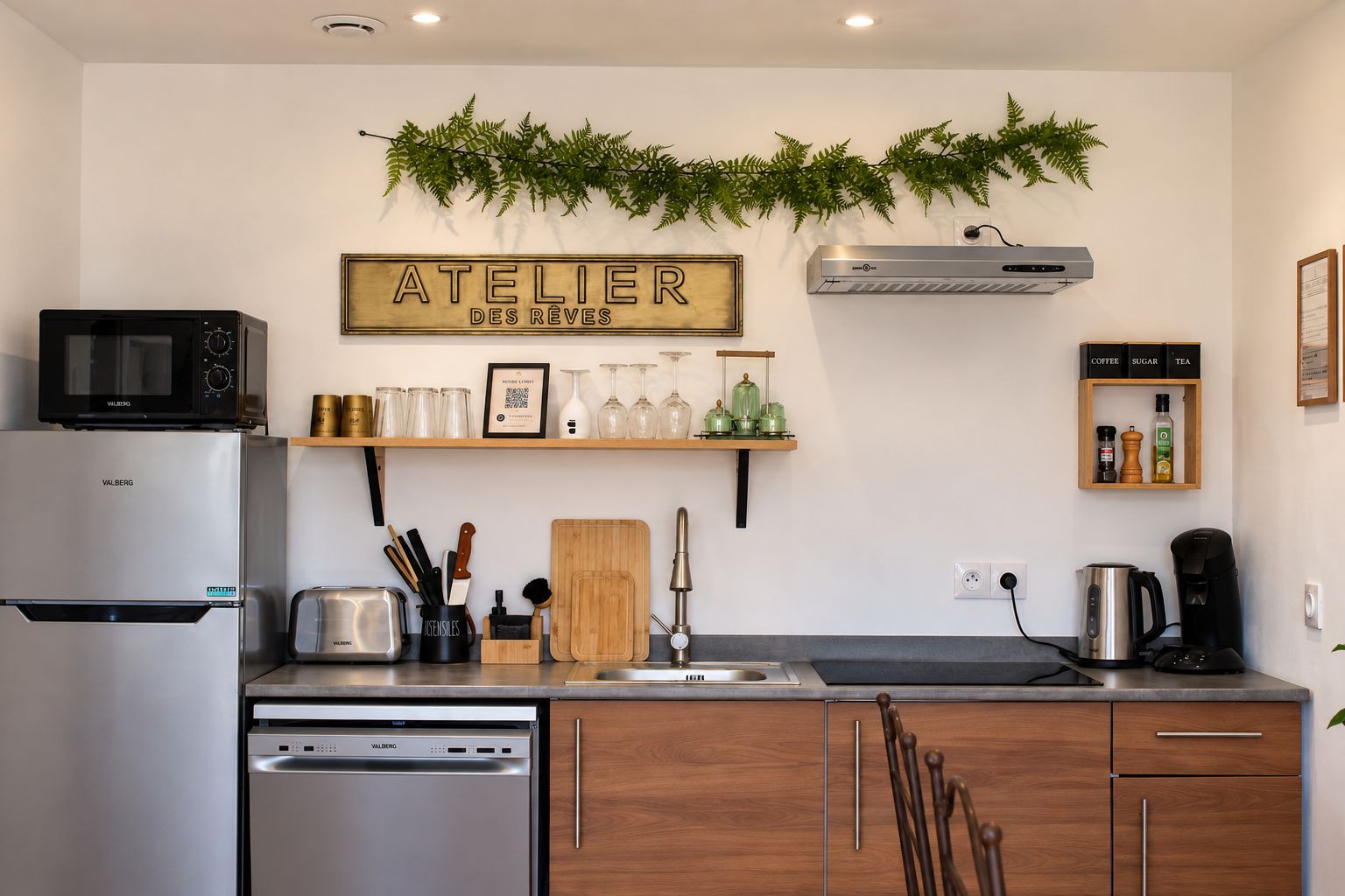 A compact kitchen with a Valberg refrigerator, a microwave, a toaster, and a dishwasher. There is a wooden cutting board, a sink, and a shelf with kitchen utensils and a framed QR code.