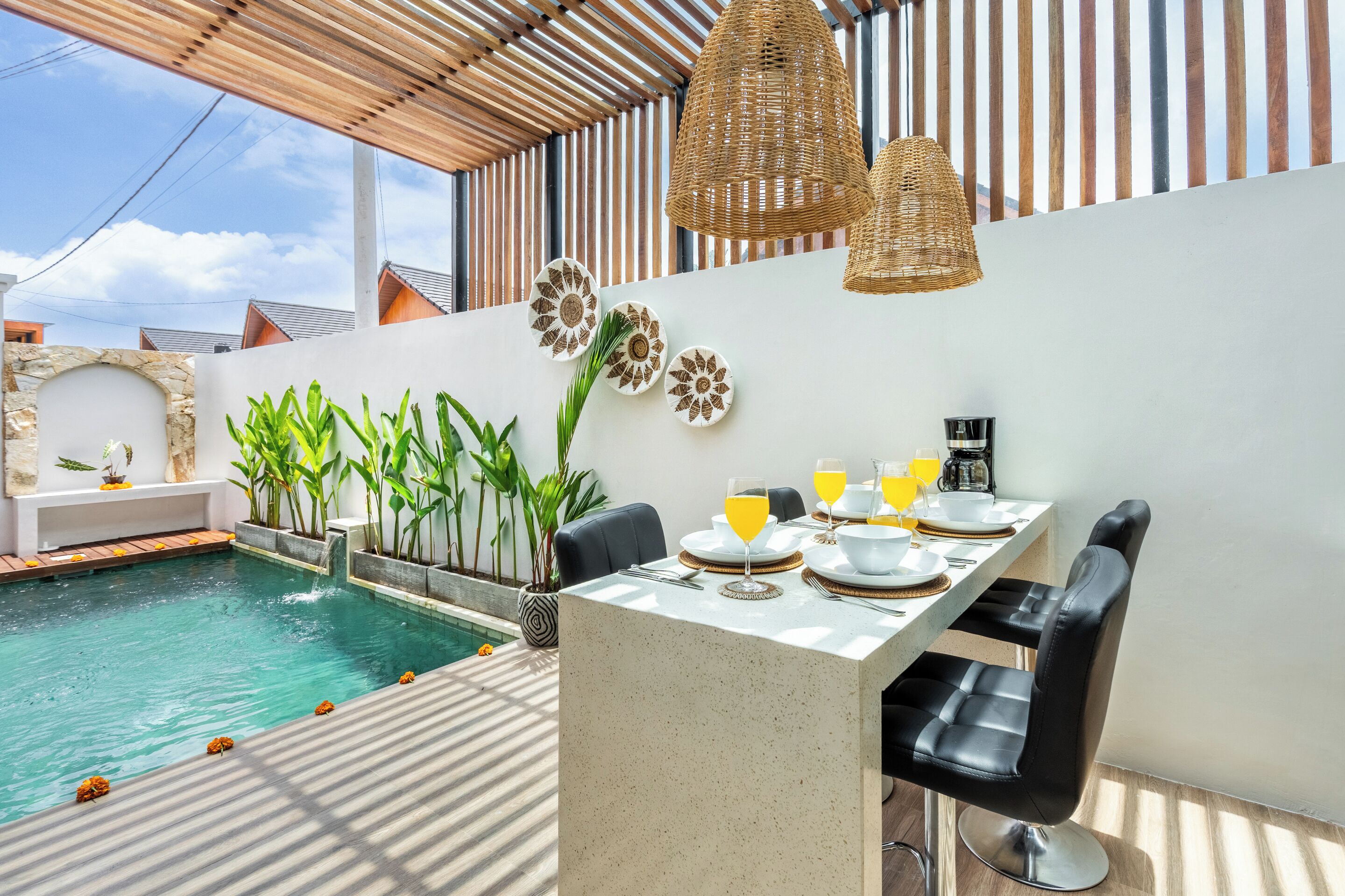 Un área de comedor junto a la piscina con una mesa para dos, rodeada de plantas en macetas, y vistas a la piscina.