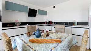 A modern kitchen with a dining area, featuring a flat-screen TV, wicker chairs, and a table set with a baguette and condiments. - Ty Glas Guesthouse – Ground Floor Room (Pleubian)