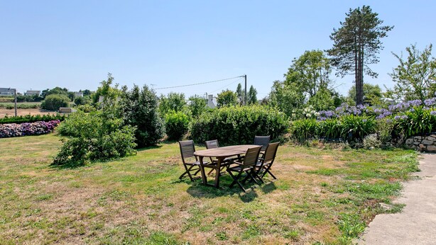 An outdoor dining area with a wooden table and chairs, surrounded by greenery and flowering plants. - Ty Glas Guesthouse – Ground Floor Room (Pleubian)