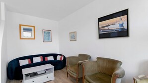 A living room with a dark blue sofa, two armchairs, a white TV stand, and framed pictures on the wall. - Ty Glas Guesthouse – Ground Floor Room (Pleubian)