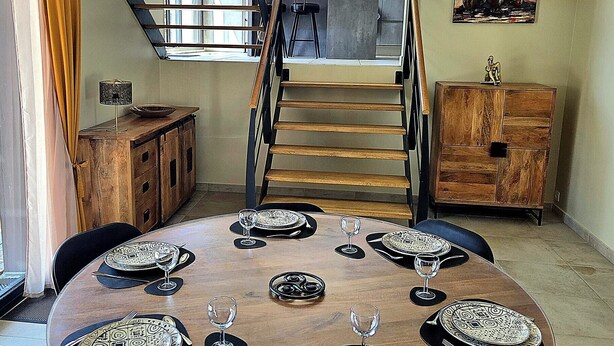 Un coin repas avec une table ronde dressée pour six personnes, un escalier et une vue sur l’extérieur.