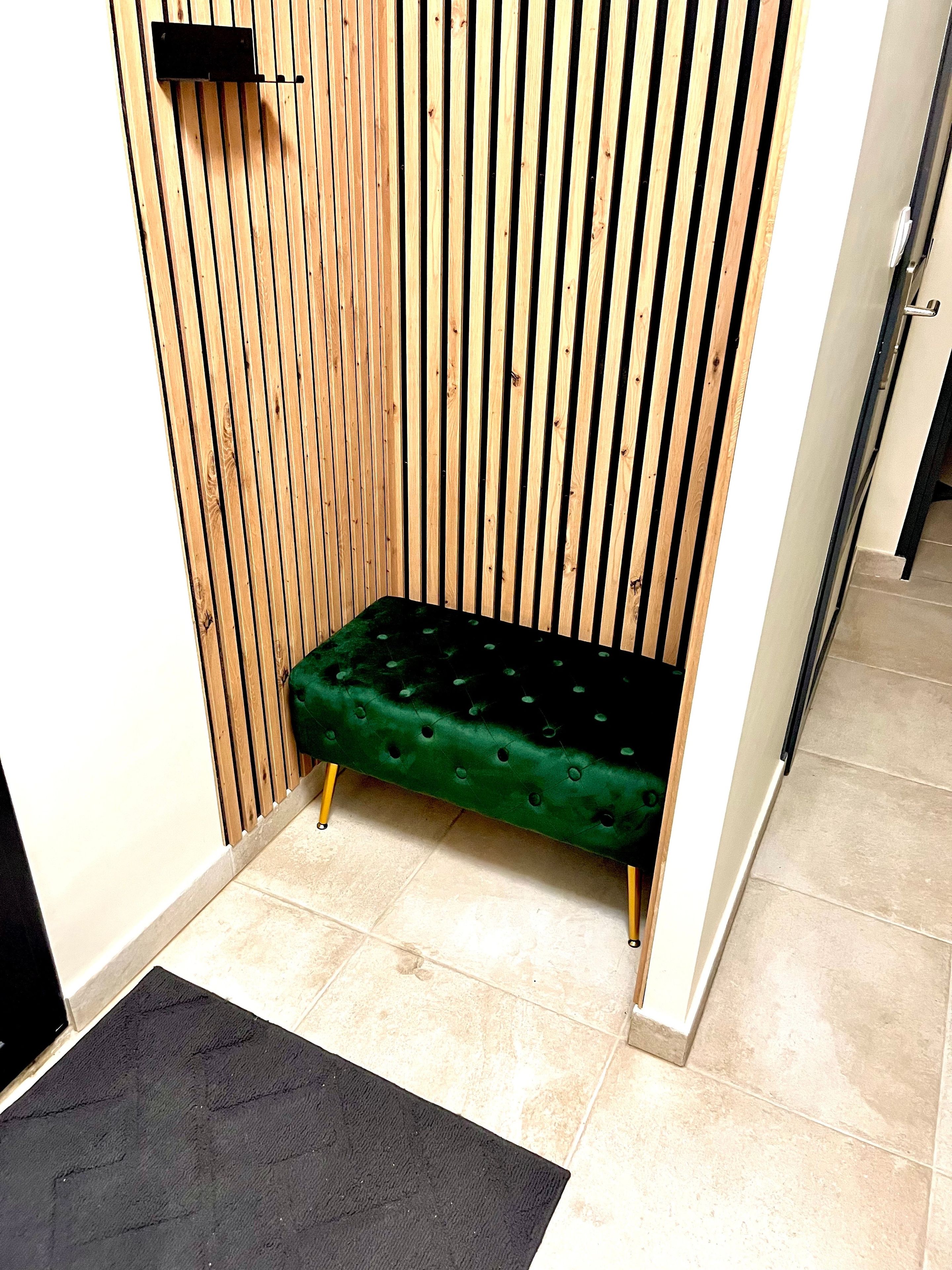 Un pequeño espacio de descanso con un banco tapizado de verde, un felpudo negro en la entrada y un muro de listones de madera.