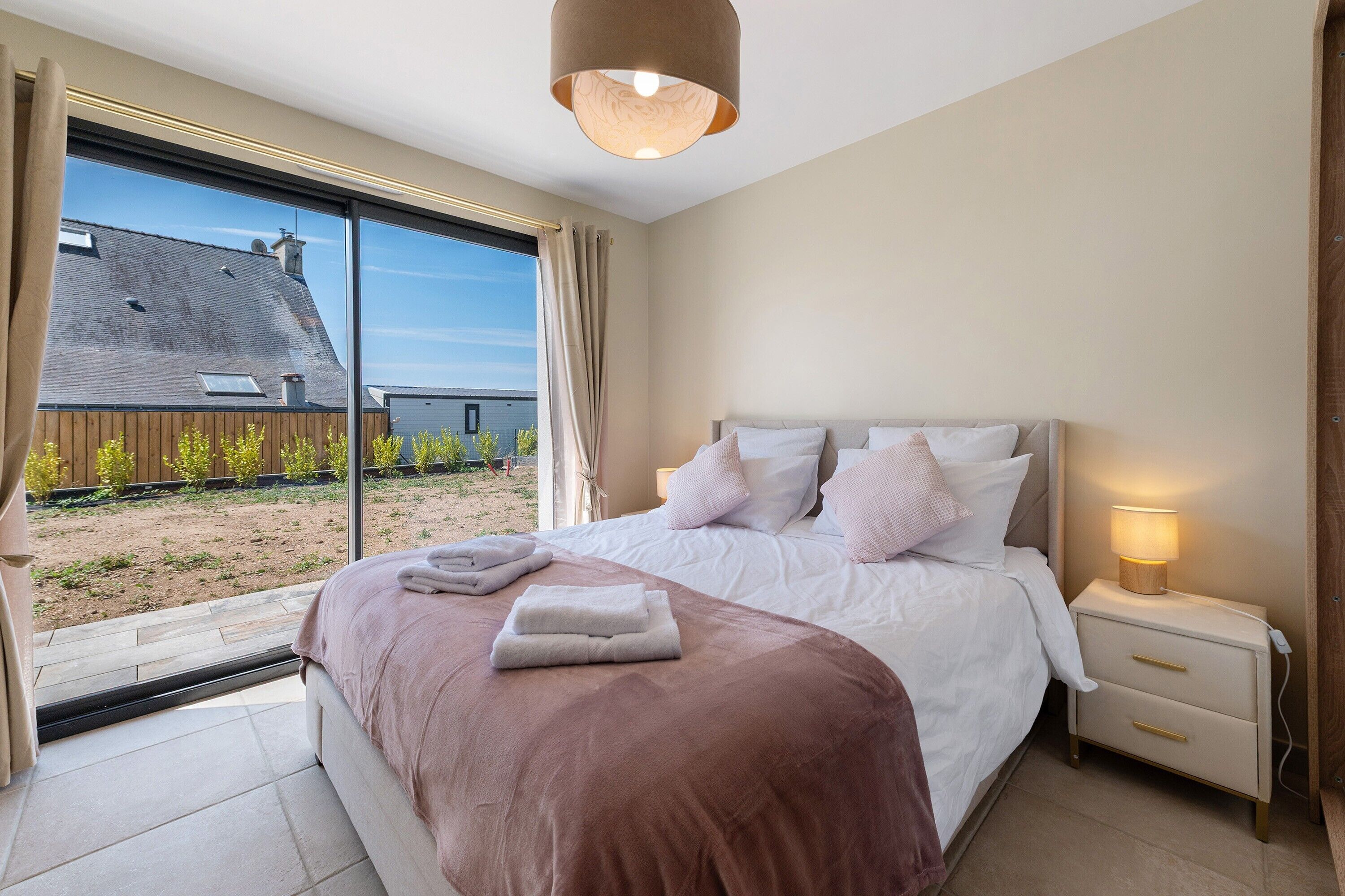A bedroom with a large bed, bedside tables, a nightstand, and a window with a view of a house and garden.