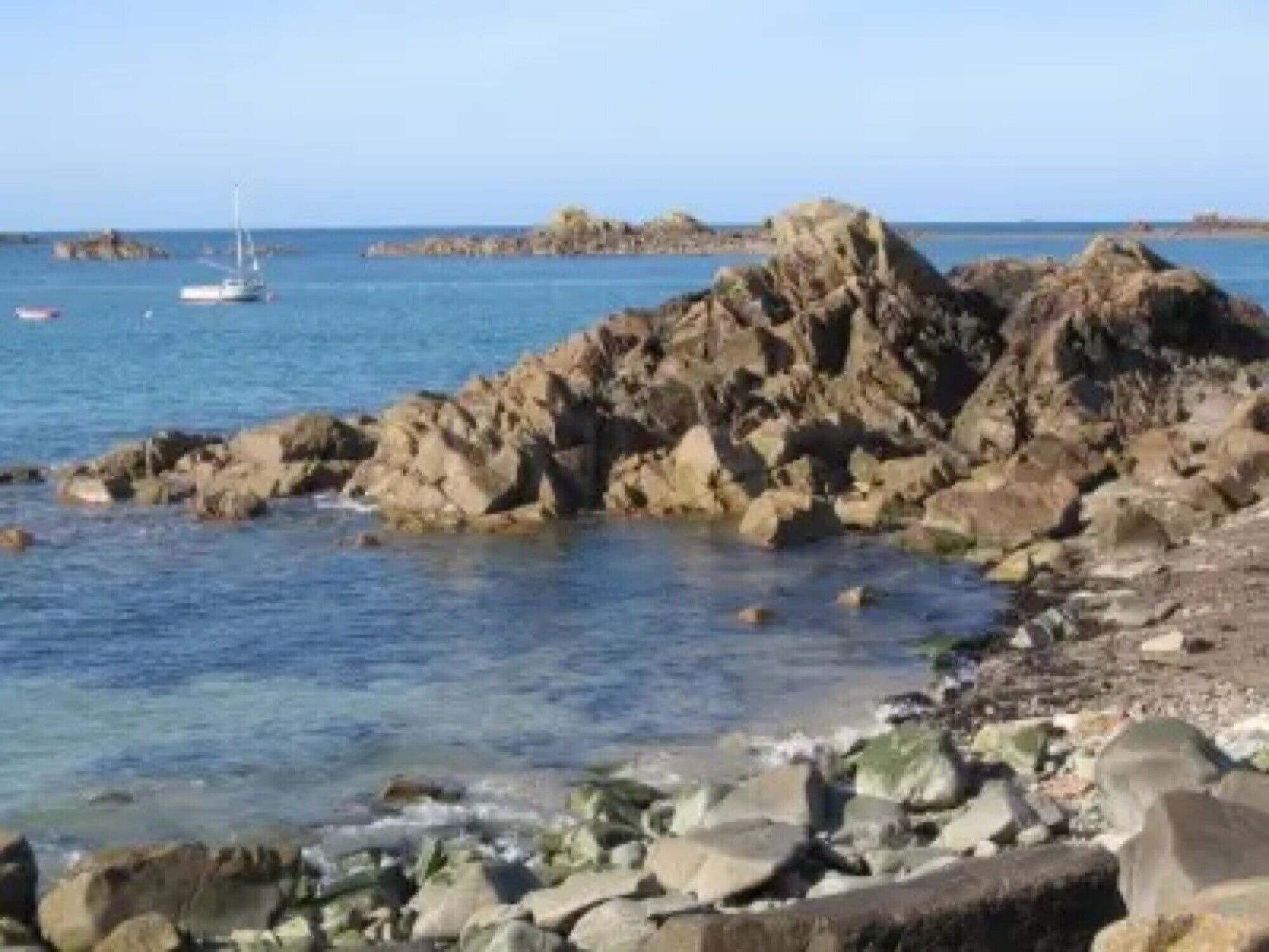 Eine felsige Küstenlinie mit klarem, blauem Wasser, ein Segelboot und Land in der Ferne.