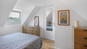 Une chambre avec un plafond en pente, un lit, une commode, une fenêtre et une porte donnant sur une autre pièce.