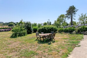 Zona pranzo all'aperto con tavolo e sedie in legno, circondata da vegetazione e piante fiorite.