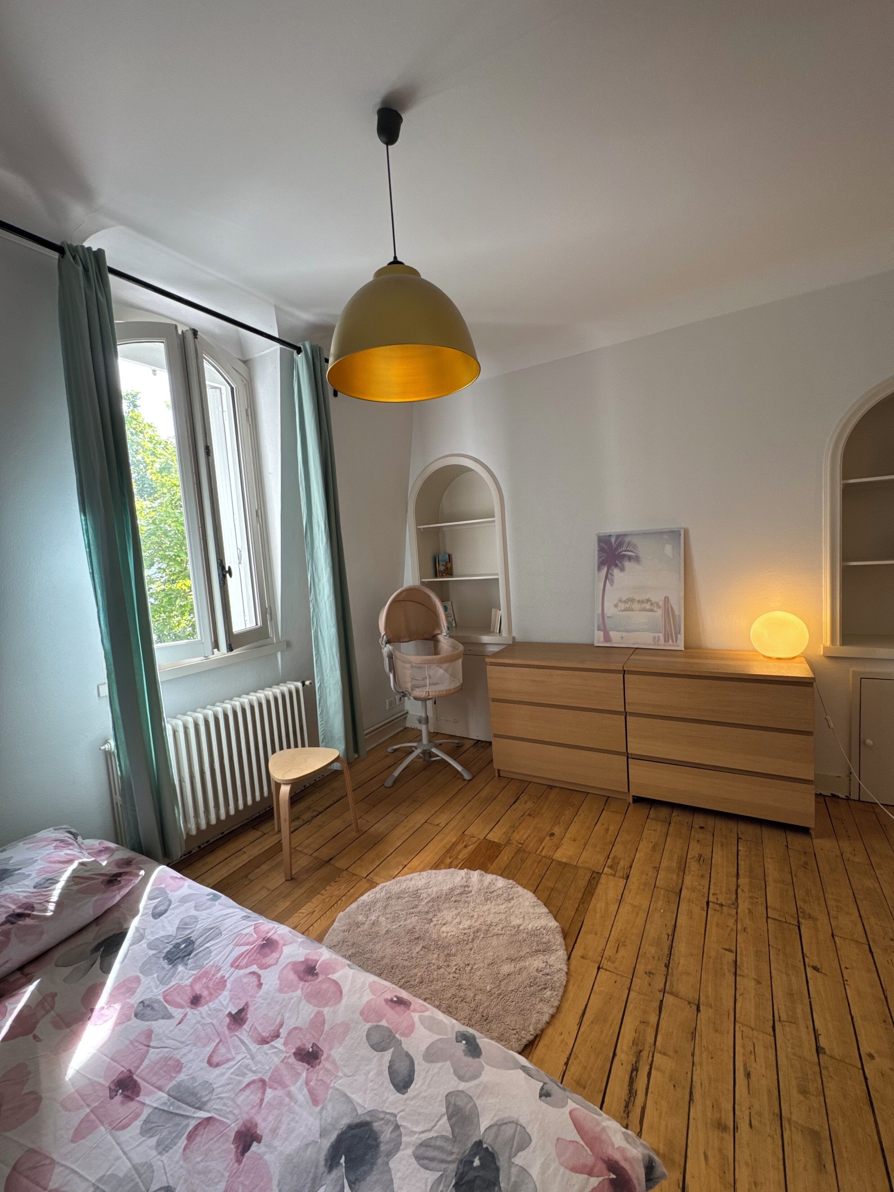 Een slaapkamer met een bed, een bureau, een stoel, een radiator en een raam met gordijnen.