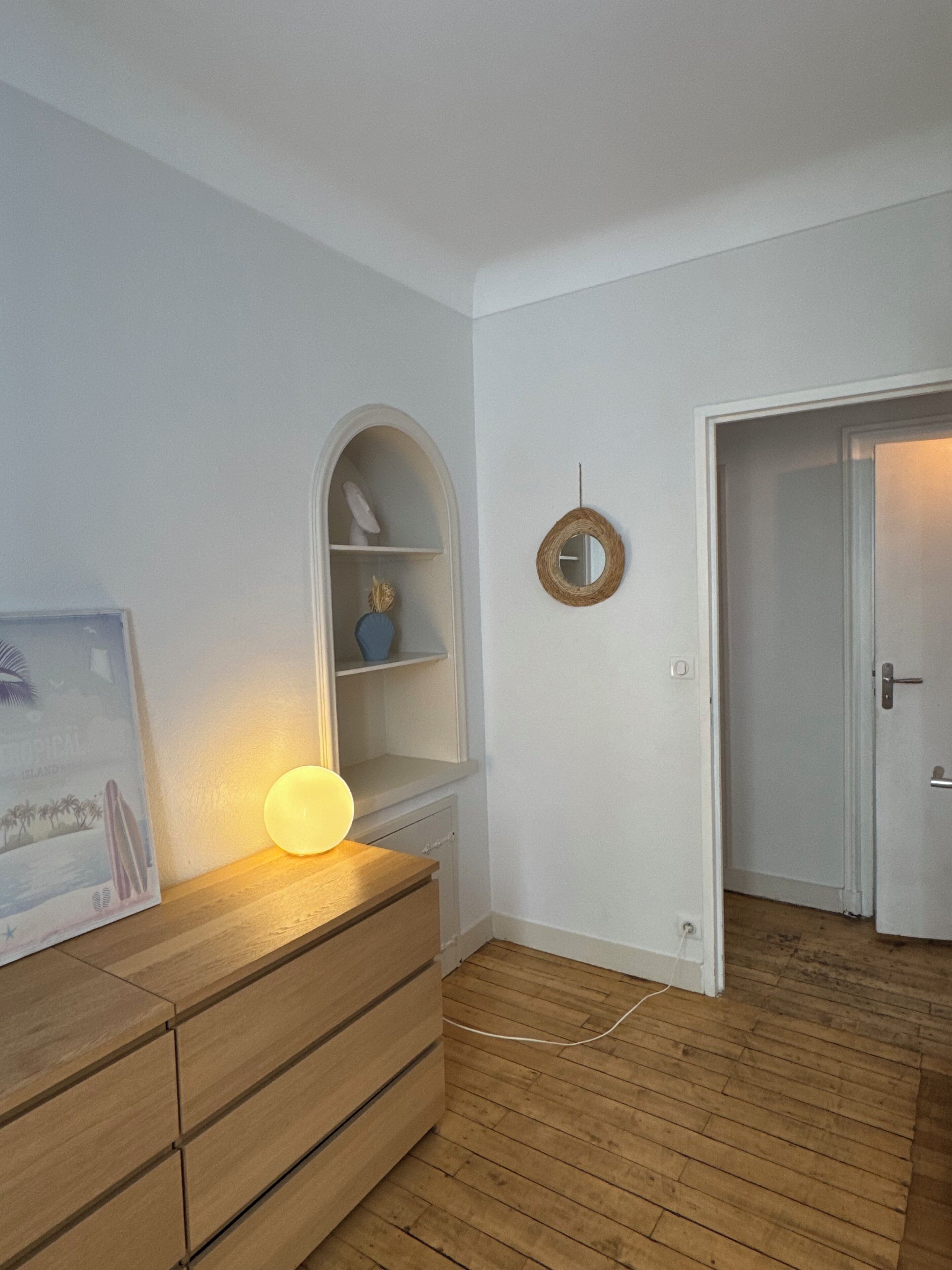 A room with a wooden dresser, a shelf with decorative items, a round mirror, and a door leading to another room.