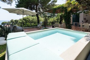 Una piscina rettangolare con bordo bianco, circondata da una recinzione in metallo nero. Sono presenti sedie a sdraio e un tavolo con sedie sotto un pergolato.